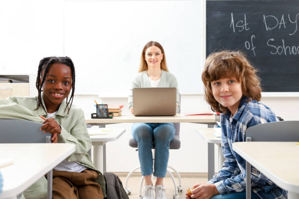 istock Smiling teacher sitting at the desk, using laptop while kids looking at the camera in classroom 2148145305