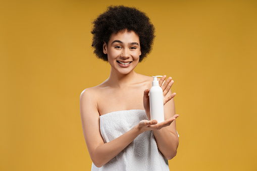 Beautiful African American young woman with curly hair in white towel after shower holding white bottle, mockup looking at camera. Advertisement concept, hygiene