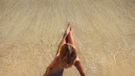 A young woman in a blue bikini sot on the sand near the waves of the blue sea. View from above.