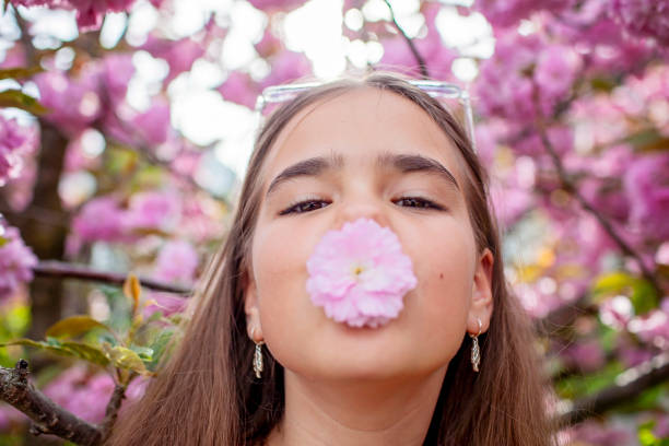 A teenage girl playfully holds a cherry blossom near her nose against a backdrop of blooming trees.