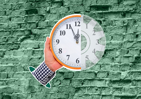 Composite Collage. Against the background of an old, weathered brick wall of pale green color, a manâs hand holds a large round clock showing the time at five minutes to twelve.