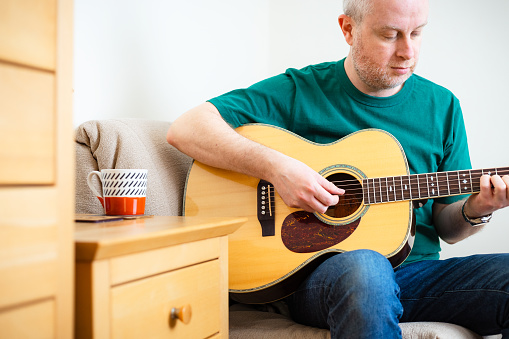 Portrait of a man in his 30s having fun and playing his acoustic guitar sitting at home. The man wears casual green t shirt and blue denim jeans.