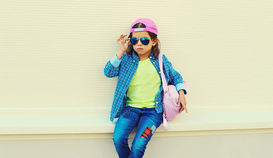 Stylish little girl child in colorful clothes posing on city street on orange wall background