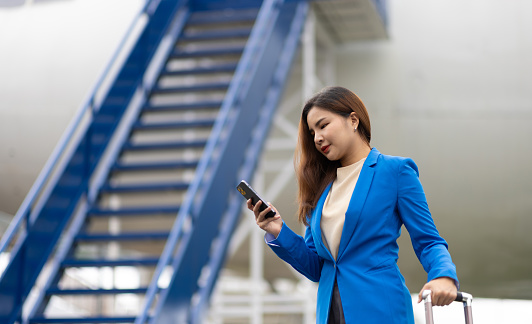 Businesswoman at the airport carries a suitcase, prepares to board the plane and depart. Asian female tourist at airport using smartphone to contact someone.