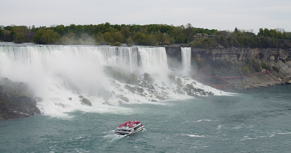 Cruise boat with tourists sails through Niagara with American Falls in background on cloudless day. Niagara cruises are must-have attraction for many tourists. Water river tourism concept.