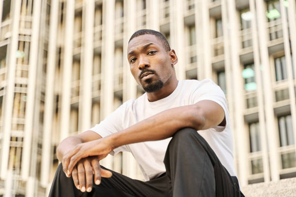 istock young black man sitting on a staircase looking at the camera seriously. he has a beard and is dressed in a plain white t-shirt. 2148159772