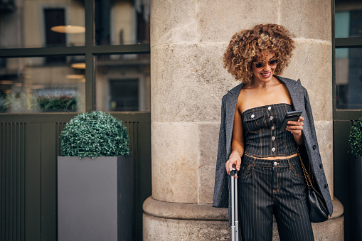 Happy young woman with curly hair texting on her phone while standing by a street with her luggage
