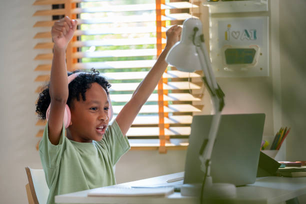 istock A cheerful boy is seated in his living room, engrossed in e-learning on his home computer. With a bright smile, he raises his hands in joy, celebrating his learning achievements. Excitement and satisfaction of a young student engaged in online education 2148162967