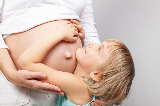 Smiling young girl embracing her pregnant mother's belly as they stand together against neutral background. Moment of family bonding and anticipation of the new baby