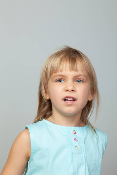 Young girl with bright blue eyes and light brown hair wearing blue dress captured gazing into distance deep in thought. Studio portrait against neutral grey backdrop