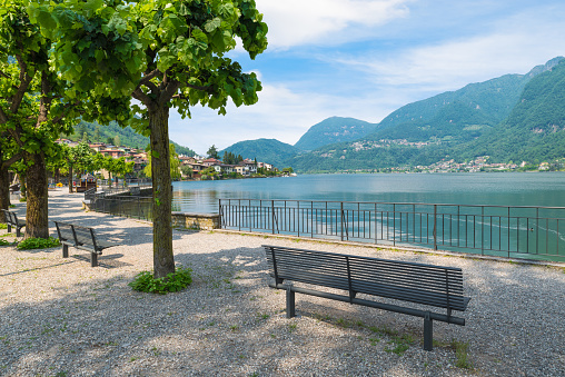 Park with benches on the shore of a lake surrounded by mountains. Lake Lugano and the town of Riva San Vitale. Riva San Vitale  is a municipality in the district of Mendrisio in the canton of Ticino in southern Switzerland, at the foot of Monte San Giorgio. The mount San Giorgio (mountain Saint George) is to be considered among the most important fossiliferous deposits in the world of the Middle Triassic and became a UNESCO World Heritage Sites in 2003