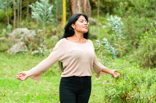 A woman stands with her arms outstretched, eyes closed, and a peaceful expression on her face. She is immersed in the tranquility of a dense forest, seemingly drawing in the fresh, soothing ambiance of her surroundings as dusk settles in.