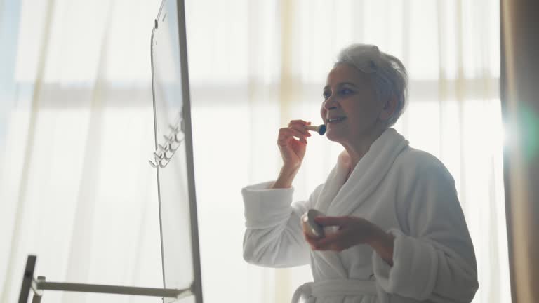 Senior woman looking in the mirror, make-up face with brush. A prosperous elderly woman looks in the mirror and applies powder to her face. Changing old age, make-up facial skin care, lifting concept.