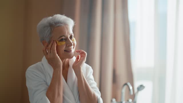 Senior woman look at mirror touch face. Wellbeing smiling woman in bathrobe looking taking care of her face apply eye patches. Getting old age change, rejuvenation face skincare self-care concept.