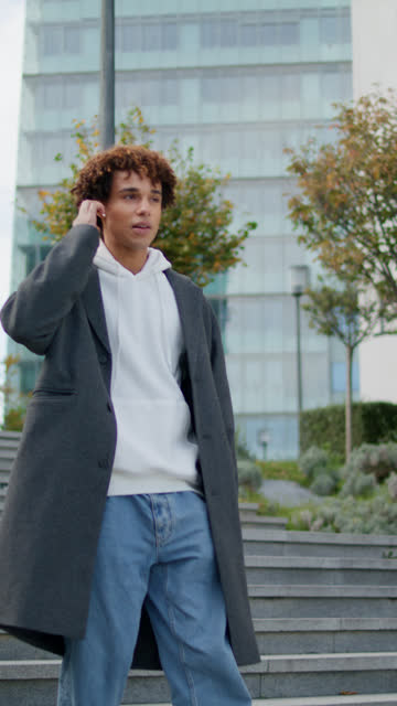 Relaxed teenager walking stairs at city street. Vertical man calling earphones