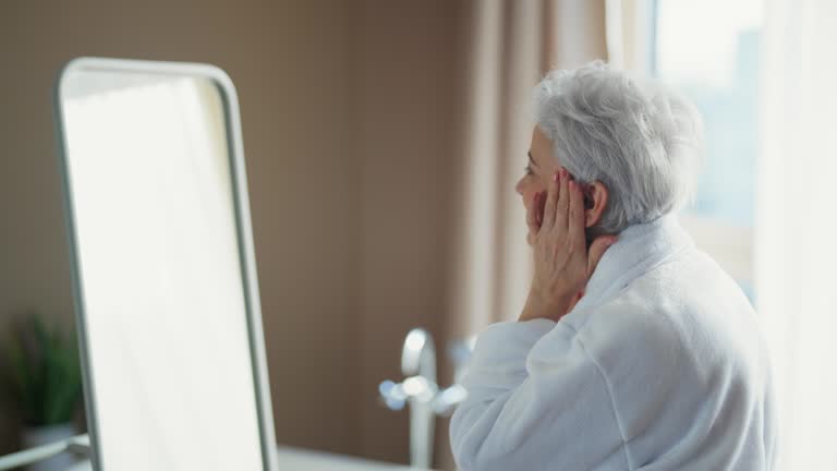 Senior woman straightens hair at home. Healthy woman in bathrobe takes care of her hair and face. Morning female routine, face skincare self-care, natural beauty aging and getting old concept.