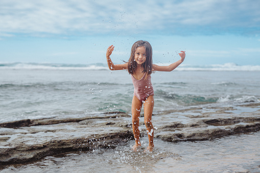 A cute preschool age girl of Pacific Islander descent wearing a swimsuit stands in the shallow ocean, splashing in the water while enjoying the beach in Hawaii.