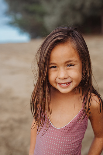 An adorable preschool age girl of Pacific Islander descent smiles at the camera while standing in the sand at the beach in Hawaii.