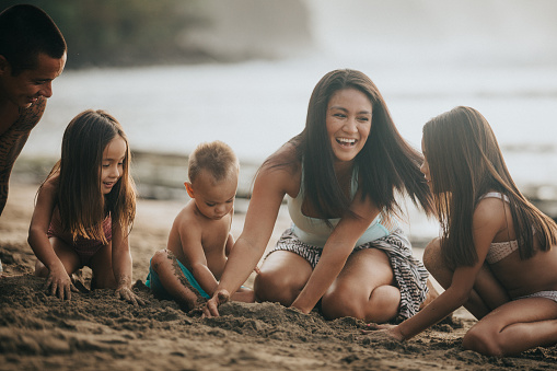 A loving and affectionate Hawaiian couple and their young children sit in the sand, building sand castles together during a beach day in Hawaii.