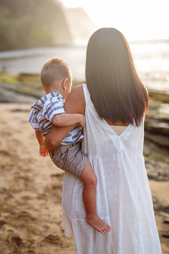 Rear view of a mother of Pacific Islander descent holds her toddler son while walking along the shoreline during a family beach day in Kauai.
