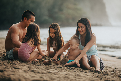 Three young Pacific Islander children sit in the sand with their mom and dad, playing in the sand together while spending the day at the beach in Hawaii.