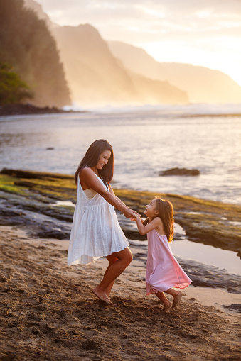 An adorable preschool age girl of Pacific Islander descent holds hands with her mother, dancing on the beach during a beautiful sunset. The two are enjoying an evening in Kauai, Hawaii together.