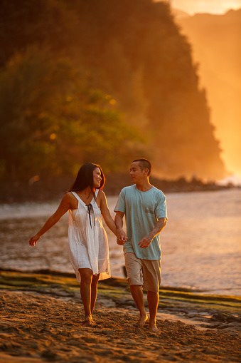 A loving and affectionate man of Pacific Islander descent holds hands with his wife while walking on the beach during a beautiful orange sunset.