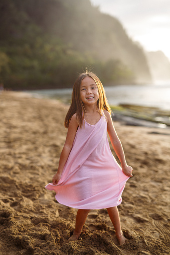 An adorable school aged girl of Pacific Islander descent holds out her dress and smiles to the camera while enjoying an evening on the beach in Hawaii.