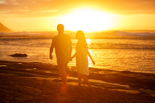 Rear view of a couple of Pacific Islander descent holding hands and walking along the beach during a beautiful sunset in Hawaii.