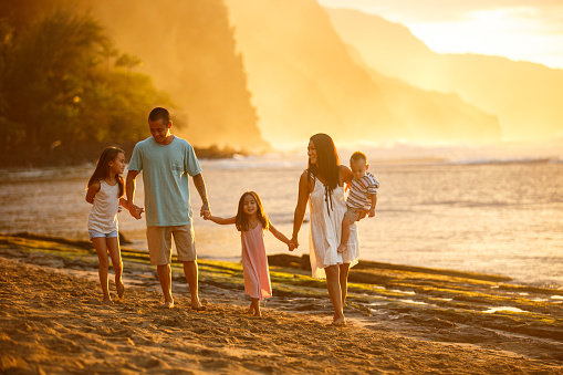 A beautiful family consisting of a Pacific Islander couple and their three young children walk on the beach together at sunset on a beautiful Winter day in Kauai, Hawaii.