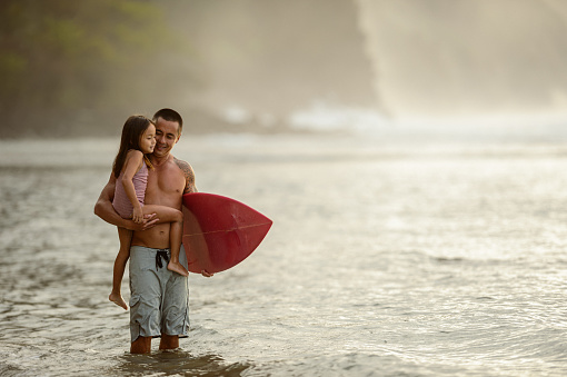 A loving and fit father of Pacific Islander descent wades in the ocean and carries his adorable preschool age girl and a surfboard while while teaching his daughter to surf.