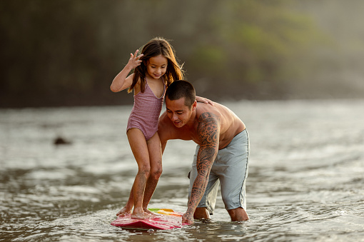 A loving father of Pacific Islander descent wades in the water while at the beach with his family and teaches his preschool aged daughter how to surf.