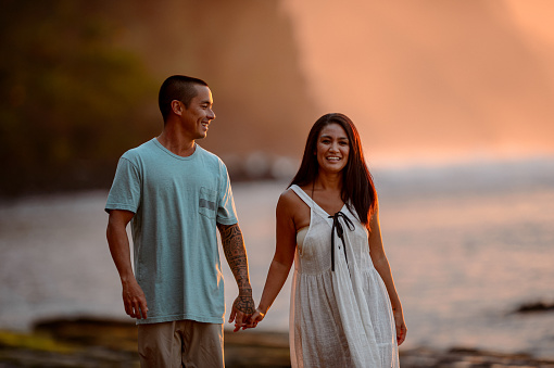 A beautiful woman of Hawaiian ethnicity smiles to the camera while holding hands with her husband and walking on the beach at sunset.