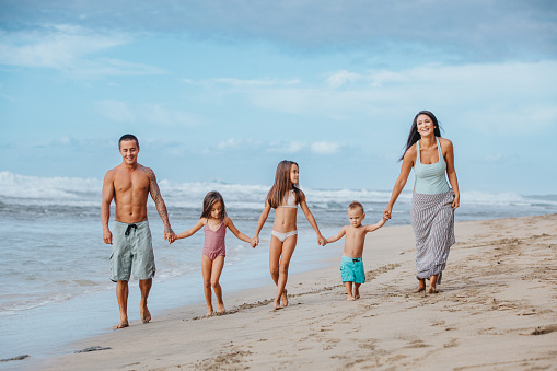 A loving family of five of  Pacific Islander descent holds each other's hands as they playfully walk down the beach together and talking happily.