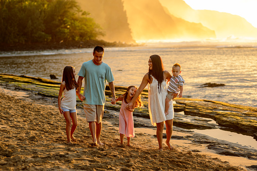 A beautiful family consisting of a Pacific Islander couple and their school age children walk on the beach together at sunset in Kauai, Hawaii.