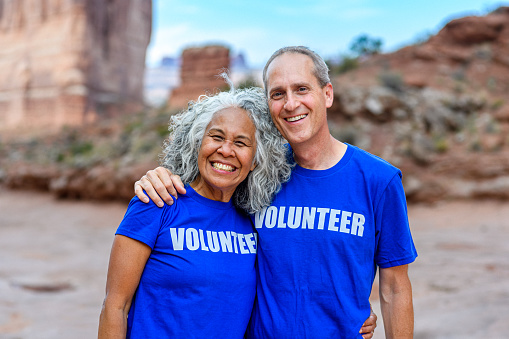 A biracial senior couple embraces and smiles to the camera while volunteering on a beautiful day during an outdoor event.