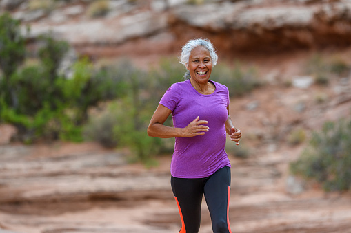 A fit and active senior woman of Pacific Islander descent smiles while trail running outside on a beautiful summer day in Utah.