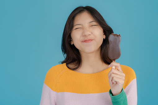 Beautiful young woman smiling happily while holding and eating ice cream in her hand on a blue background