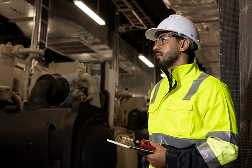 Smart HVAC system engineer with a green safety jacket and PPE holds a digital tablet and walkie-talkie working at the site line of the HVAC control room