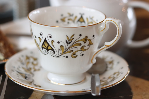 A patterned China teacup and saucer on a table in front of a defocussed teapot
