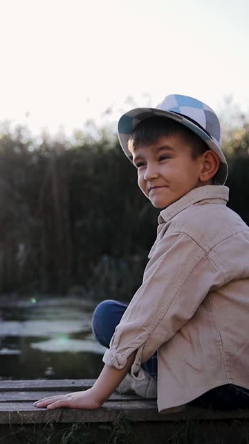 Boy near pond or river spending his summer holidays time in countryside. Small preschool boy is looking to the camera and smile. Vertical video