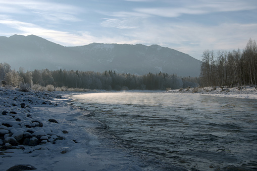 A frozen river surrounded by snow-covered mountains under a cloudy sky. The natural landscape features a sloping terrain and a freezing cold atmosphere. Russia, Chuisky ridge, Katun river.