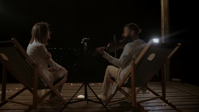 A man plays the violin and a girl sings at a concert on the street at night. A duo of musicians plays under the night sky. Musicians play on the stage of the restaurant.