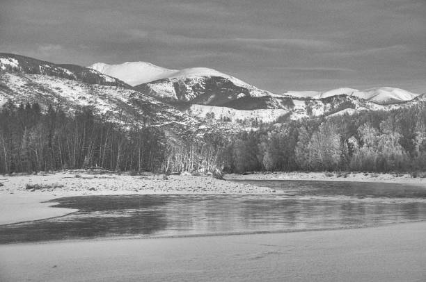 A frozen river surrounded by snow-covered mountains under a cloudy sky. The natural landscape features a sloping terrain and a freezing cold atmosphere. Russia, Chuisky ridge, Katun river.