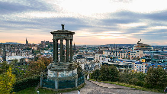 Aerial view of Calton Hill, Edinburgh sunset aerial view, Gothic Revival architecture in Scotland, View of Edinburgh city center from Calton Hill\n\nCalton Hill is a hill in central Edinburgh, Scotland, situated beyond the east end of Princes Street and included in the city's UNESCO World Heritage Site. Views of, and from, the hill are often used in photographs and paintings of the city.\nCalton Hill is the headquarters of the Scottish Government, which is based at St Andrew's House, on the steep southern slope of the hill. The Scottish Parliament Building and other prominent buildings such as Holyrood Palace lie near the foot of the hill. Calton Hill is also the location of several monuments and buildings: the National Monument, the Nelson Monument, the Dugald Stewart Monument, the old Royal High School, the Robert Burns Monument, the Political Martyrs' Monument and the City Observatory.\nThe area lies between the Edinburgh districts of Greenside and Abbeyhill.\n\nEdinburgh is the capital city of Scotland and one of its 32 council areas. The city is located in south-east Scotland, and is bounded to the north by the Firth of Forth estuary and to the south by the Pentland Hills. Edinburgh had a population of 506,520 in mid-2020, making it the second-most populous city in Scotland and the seventh-most populous in the United Kingdom.