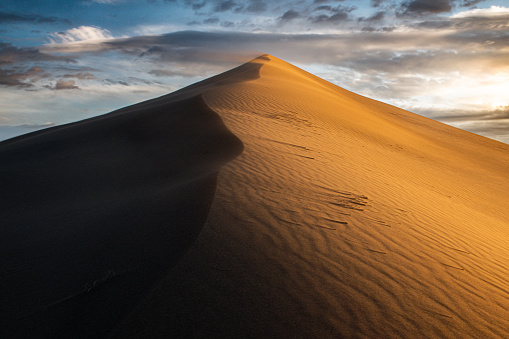 Glowing with the warm light of the rising sun, a sand dune rises sharply against a cloudy sky.
