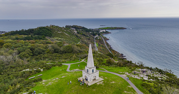 Aerial view of the Obelisk on Killiney Hill in Wicklow-Ireland, Killiney Obelisk, popular tourist destinations in ireland, people enjoy the view next to the monument on the hill, view hill\n\nThe Killiney Obelisk was built in 1742 to commemorate what is described as ‘the Year of the Slaughter’ or Ireland’s forgotten famine. In 1740 there was an incredibly cold winter which was followed by a severe summer of rain and floods. This wiped out the crops and killed off livestock. The following winter was again BALTIC with temperatures not getting above minus 10 for a month. It was so cold that small vessels were destroyed by icebergs on the River Liffey, street lamps could not be lit plunging towns and villages into darkness and food riots were common throughout the land. Many of the oldest trees were also felled for fuel and sickness was common all over. Reports suggest that up to 480,000 may have people died during this forgotten famine.  A small number of rich landlords like John Malpas of Killiney Hill and Kathryn Connolly of Castletown House commissioned famine relief projects like this one to provide employment to destitute families. Ironically this monument now overlooks the wealthiest part of Ireland.