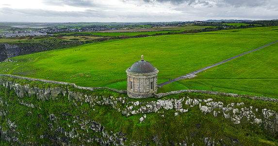 Aerial view of Mussenden Temple library in Northern Ireland, Downhill, County Londonderry, Ruin of Mussenden temple on the edge of a cliff, Aerial view of Causeway Beach and Mussenden Temple, Mussenden Temple on high cliffs near Castlerock\n\nMussenden Temple is located in the beautiful surroundings of Downhill Demesne near Castlerock in County Londonderry. It perches dramatically on a 120 ft cliff top, high above the Atlantic Ocean on the north-western coast of Northern Ireland, offering spectacular views westwards over Downhill Strand towards Magilligan Point and County Donegal and to the east Castlerock beach towards Portstewart, Portrush and Fair Head.