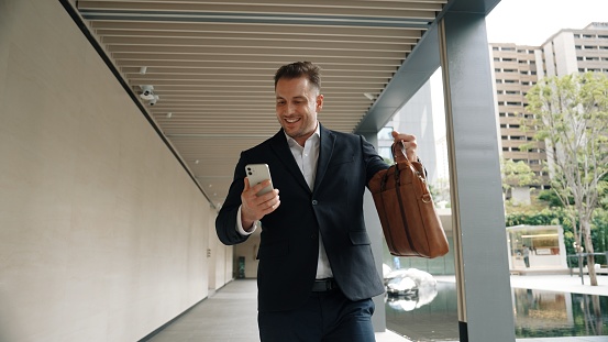 Happy project manager checking mobile phone while walking at corridor. Professional business man celebrate successful project or increasing sales while wear suit and hold suitcase for meeting. Urbane.