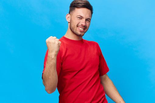 Overjoyed happy tanned handsome man in red t-shirt raise fist up have big win say Yeah posing isolated on blue studio background. Copy space Banner Mockup. People emotions Lifestyle concept